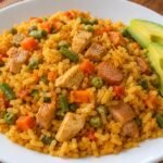 Arroz mixto colombiano casero con pollo, cerdo, salchichas, zanahoria, habichuelas y arvejas, acompañado de aguacate en un plato blanco.