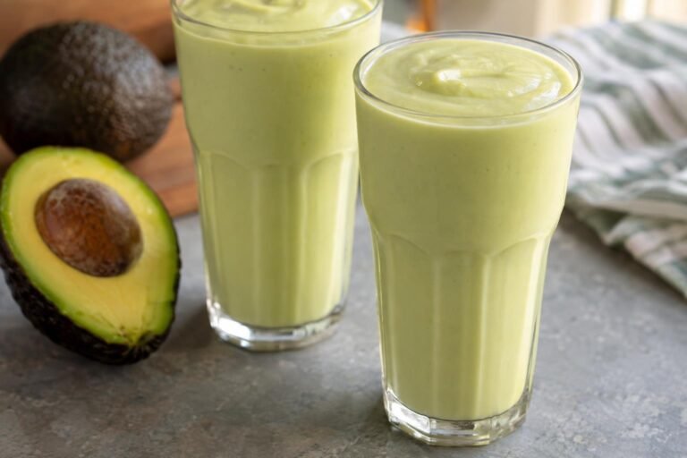 Dos vasos de batido de aguacate brasileño cremoso, preparado con leche, azúcar y hielo, junto a medio aguacate maduro.