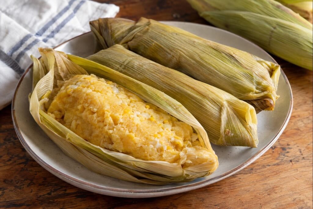Tres envueltos de mazorca colombianos cocidos al vapor, uno abierto para mostrar su masa amarilla y húmeda dentro de la hoja.