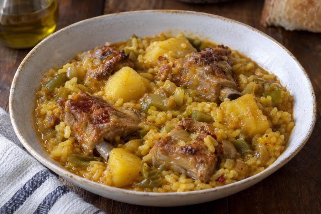 Plato hondo de patatas con costilla de cerdo y arroz en salsa espesa dorada, con cebolla y pimiento visibles.