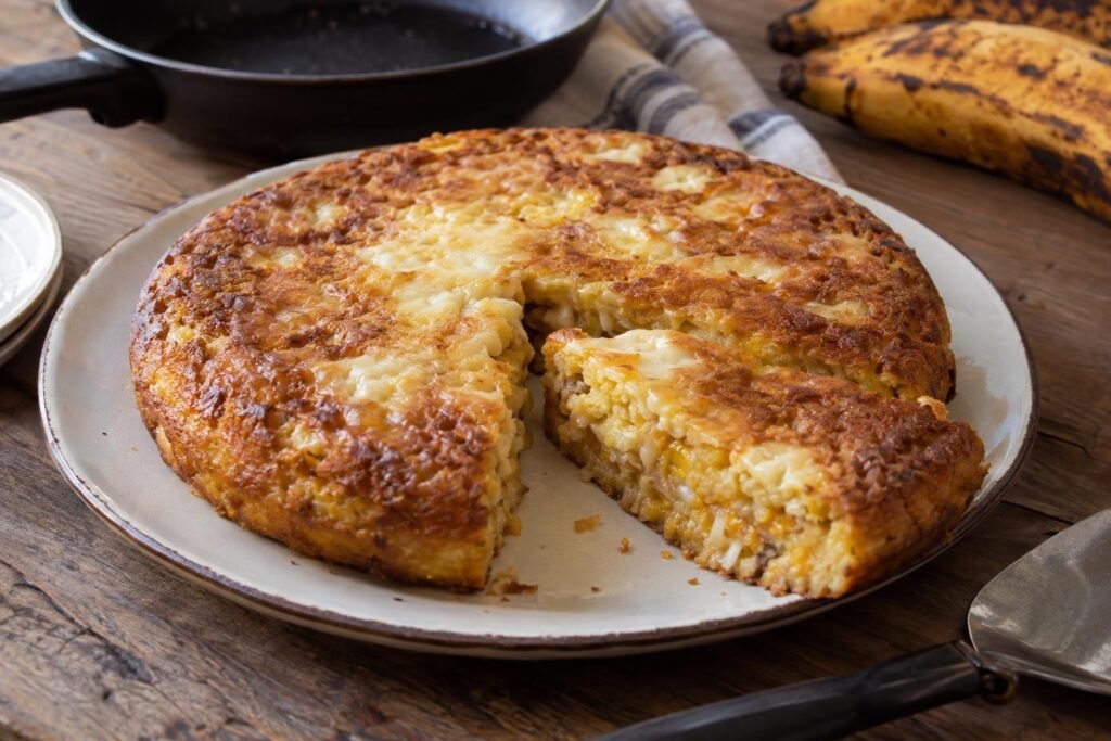 Torta redonda de plátano maduro con queso, dorada y cortada en porciones, preparada en sartén.